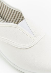 Zapatilla blanca de lienzo sin cordones con un upper de tela texturizada y paneles elásticos a los lados. El interior cuenta con un forro a rayas. Diseño minimalista.