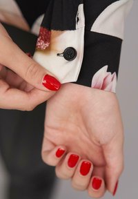 Zwarte bloemen lange mouw shirtmanchet met een zwarte knop, gladde stof, en een huidkleurige hand met rode nagellak die de manchet aanpast.
