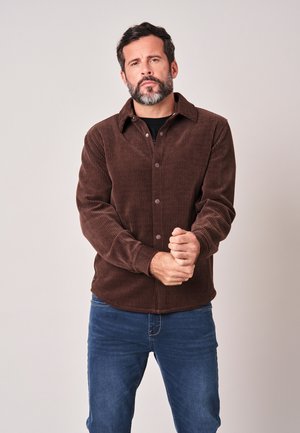 Man met baard die een bruine ribcordjas en blauwe spijkerbroek draagt, staat met de handen gevouwen voor een effen lichte achtergrond.