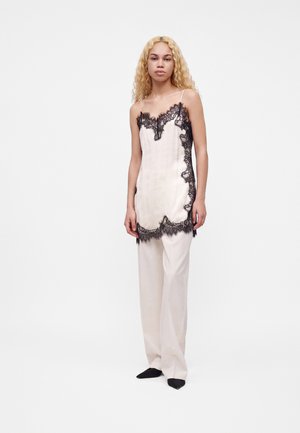 Femme aux longs cheveux blonds bouclés, portant un haut en satin blanc avec dentelle noire et un pantalon large assorti, debout devant un fond blanc.
