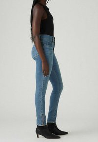 Vaqueros de mezclilla de tiro alto en azul, con corte ajustado, detalles de cierre lateral en los tobillos, combinados con botas negras hasta el tobillo.