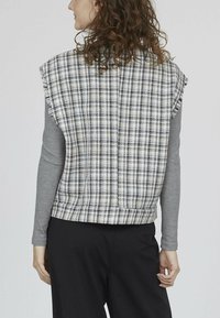 Grå langærmet skjorte under en cropped ærmeløs ternet vest i sort, hvid og gul med frynsede kanter og en lige kant.