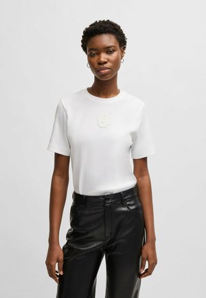 T-shirt bianco a maniche corte con un design floreale testurizzato bianco sul petto, abbinato a pantaloni di pelle neri. Forma semplice e aderente.