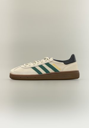 Baskets Adidas Spezial blanches avec des rayures vertes, semelle marron, languette de talon noire et logo jaune sur le côté, présentées de profil.