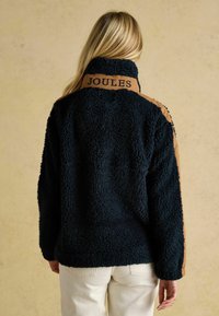 Granatowa kurtka polarowa z wysokim kołnierzem i brązowymi, kontrastującymi panelami na ramionach, z logo "JOULES" na plecach.