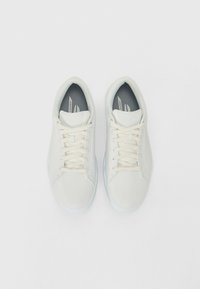 Baskets blanches avec une finition lisse et mate. Présentent des lacets plats, une languette texturée et une semelle translucide bleu clair. Design minimaliste.