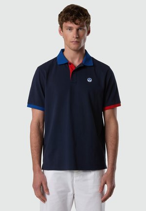 Giovane uomo con capelli corti e ricci, indossa una polo blu navy con dettagli sul colletto blu e rossi e pantaloni bianchi, in piedi davanti a uno sfondo neutro.