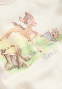 Cremefarvet stof viser et grafisk motiv af en hjort og en fugl, der leger omkring en træstub, med ordet "Bambi" broderet i lyseblå.