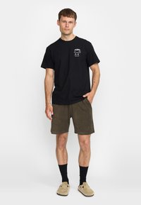 Zwarte katoenen t-shirt met een kleine witte graphic op de borst, gecombineerd met olijfgroene shorts. Het model draagt zwarte sokken en beige instapschoenen.
