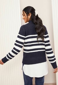 Marineblau- und weiß gestreifter Pullover mit hochgeschlossenem Kragen, gerippten Bündchen und Saum, aus weichem Material, über einem weißen Hemd getragen.