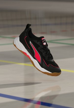 Zapatilla deportiva Puma negra y roja con suela blanca y dibujo de goma, mostrada en el aire sobre una superficie lisa de pista cubierta con líneas de colores.