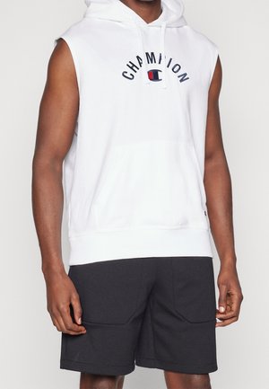 Hoodie sans manches blanc de Champion avec cordons de serrage et poche avant, porté avec un short noir doté de grandes poches avant.