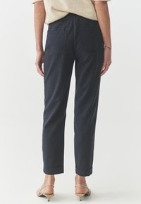 Navy blue trousers with a soft texture, featuring a relaxed fit, cuffed hems, and two back pockets. Worn with ankle strap shoes.