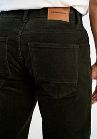 Pantalones de pana verde oscuro con una superficie texturizada, que cuentan con dos bolsillos traseros de parche y una etiqueta de marca de cuero en la cintura.