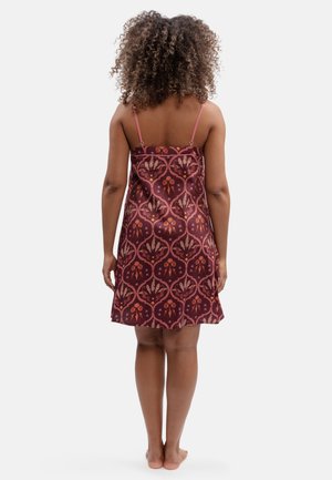 Mujer con cabello rizado, de pie descalza de espaldas, vistiendo un vestido sin mangas burdeos con un patrón floral.