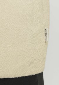 Beige szövet, struktúrált felülettel, fekete alsó résszel. Kis címke található rajta, rajta a "JACK & JONES" felirattal.