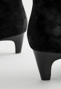Czarne zamszowe botki za kostkę o eleganckim wzornictwie, z odkrytą, spiczastą końcówką i subtelnym połyskiem. Posiadają zwężający się czarny obcas dla dodatkowej elegancji.