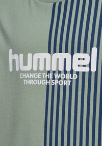 Chemise verte à manches courtes avec des rayures verticales marines. Présente un grand texte blanc "hummel" et "CHANGE THE WORLD THROUGH SPORT" en dessous.