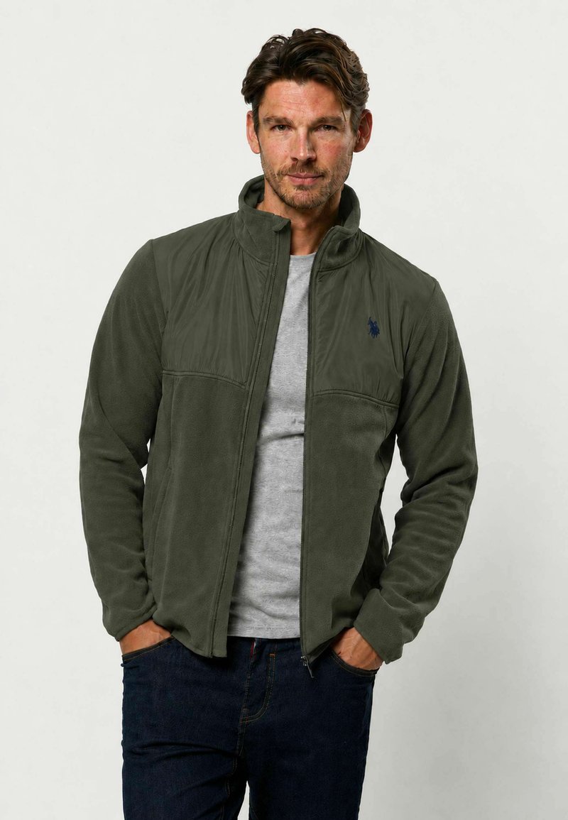 Homme portant une veste polaire verte zippée par-dessus un t-shirt gris, les mains dans les poches d'un jean bleu foncé, sur fond uni.