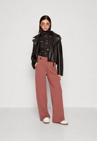 Jovem mulher vestindo uma jaqueta de couro preta, um suéter de gola alta com padrão celestial preto, calças largas cor-de-rosa, sapatos brancos e óculos de sol com tonalidade laranja.