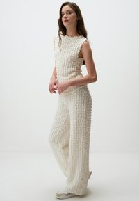 Weißes, strukturiertes Zweiteiler-Outfit mit einem ärmellosen Oberteil und weit geschnittenen Hosen, gefertigt aus einem gemusterten Stoff und einem subtilen Lagen-Design.