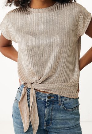 Vrouw draagt een metalen geribd kortermig shirt met een knoop aan de voorkant, gecombineerd met blauwe spijkerbroek, handen rustend op de heupen.