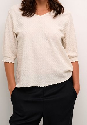 Femme portant un haut crème texturé à manches trois-quarts et un pantalon noir, les mains dans les poches, sur un fond clair uni.