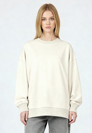 Jonge vrouw met blond haar draagt een oversized crèmekleurige trui en grijze jeans tegen een effen lichte achtergrond.