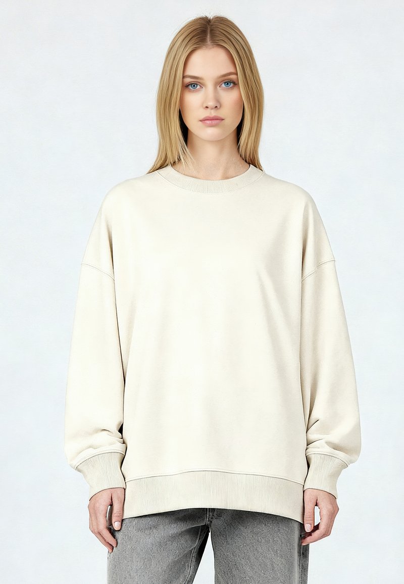 Jeune femme aux cheveux blonds portant un sweat-shirt crème surdimensionné et un jean gris, devant un fond clair uni.