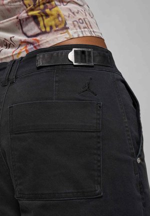 Un pantalon en denim noir présentant une poche arrière avec un logo en relief, une fermeture à boucle argentée et une finition texturée. Coupe standard avec passants de ceinture.