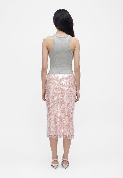 Femme aux longs cheveux foncés portant un haut sans manches gris et une jupe midi rose à sequins, debout de dos sur un fond blanc.