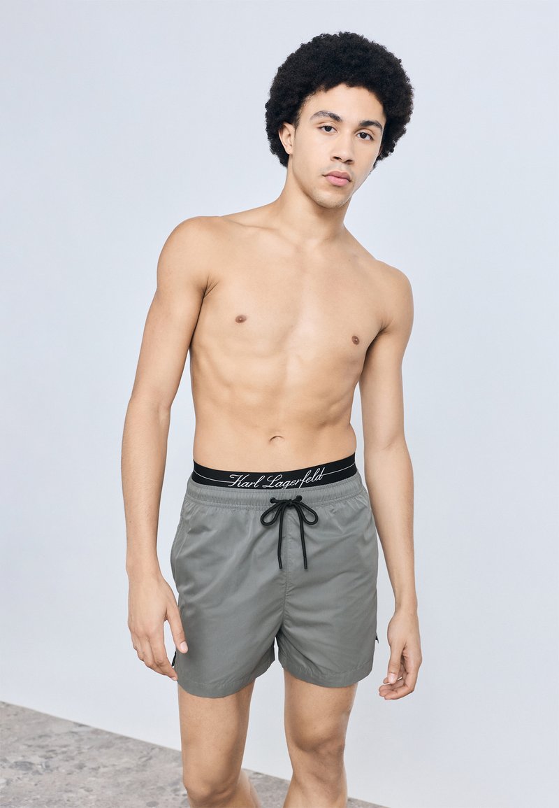 Szürke úszónadrág fekete húzózsinórral, márkázott derékpánttal. Sima textúrájú, térdig érő kialakítás. A modell felsőruházat nélkül látható.