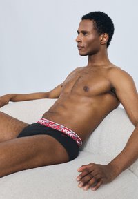 Černé boxerky s bílým a červeným vzorovaným pasem s logem Nike. Hladká tkanina, přiléhavý střih, sedící póza na světlém pozadí.