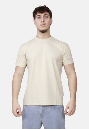 Giovane uomo con capelli corti e ricci, indossa una maglietta beige a maniche corte e jeans azzurri, in piedi davanti a uno sfondo bianco.