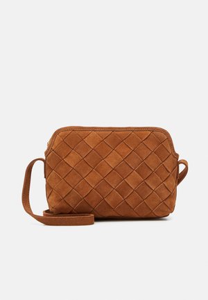 Sac bandoulière en suède marron avec un motif matelassé en losanges et une longue bandoulière sur un fond gris clair uni.