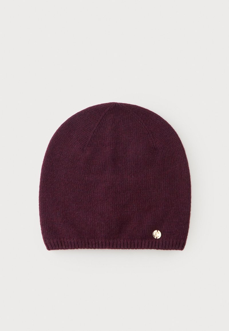 Bonnet en tricot violet foncé avec un petit emblème rond doré près du bord inférieur, présenté sur un fond clair uni.