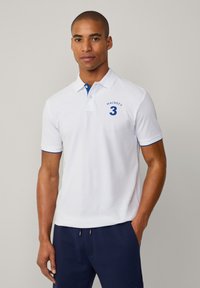 Άσπρο polo μπλουζάκι με μπλε γιακά και αχτίνες μανικιών. Διαθέτει κεντημένη την ένδειξη "HACKETT" και τον αριθμό "3" στο στήθος, κατασκευασμένο από μαλακό βαμβακερό ύφασμα.