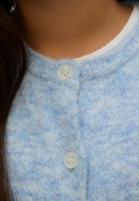 Šviesiai mėlynas megztas kardiganas su apvalia iškirpte, pasižymintis lygiu paviršiumi ir dviem baltais plastikiniais mygtukais priekyje.