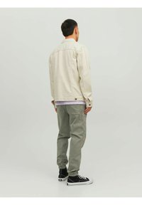 Giacca beige chiaro, tessuto di cotone, polsini con bottoni, vestibilità comoda, abbinata a pantaloni verde chiaro e sneakers nere, stile rilassato.