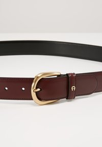 Bordeaux leren riem met een gladde textuur, voorzien van een gouden metalen gesp en witte stiksels, met een zwarte onderkant.