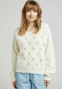Pull blanc et duveteux présentant un motif en losange agrémenté de dorures. Il a un col rond et des manches longues et volumineuses.