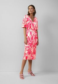 Fließendes, kurzärmliges Kleid in Pink mit floralen Mustern; V-Ausschnitt-Design, knielang. Kombiniert mit pinken Sandalen mit Absatz. Glatte Stofftextur.