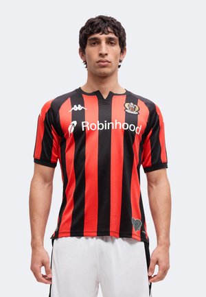 Jeune homme portant un maillot de football rayé rouge et noir avec le logo du sponsor "Robinhood" et un short blanc, se tenant devant un fond uni.