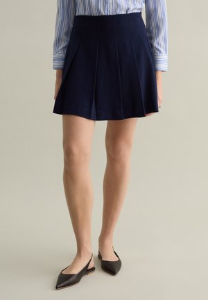 Falda plisada azul marino hecha de una tela de textura suave. Combinada con bailarinas negras con tiras en el talón, con punteras puntiagudas y un diseño minimalista.