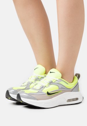 Benen dragen neongele, grijze en witte Nike-sneakers met zwarte swoosh-logo's, staande op een witte ondergrond.