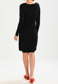 Robe noire à manches longues avec un col rond, créant une silhouette simple. Texture lisse, longueur jusqu'aux genoux, accompagnée de chaussures orange vif.