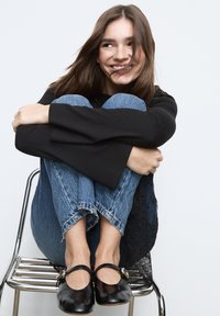 Zwart shirt met lange mouwen en uitlopende manchetten, lichtblauwe distressed jeans en zwarte Mary Jane schoenen met gouden accenten, zittend op een metalen stoel.