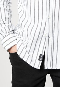 Witte shirt met zwarte verticale strepen, knopen en een label. Gecombineerd met zwarte broek, met een casual, slim-fit ontwerp.