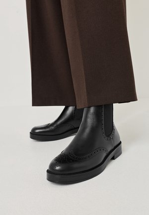 Bottines brogue en cuir noir sur une personne portant un pantalon large marron sur fond clair.