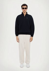 Μαύρο φλις φούτερ με φερμουάρ και ψηλό γιακά, συνδυασμένο με άνετο λευκό παντελόνι και λευκά αθλητικά παπούτσια. Αξεσουάρ περιλαμβάνουν μαύρα γυαλιά ηλίου.